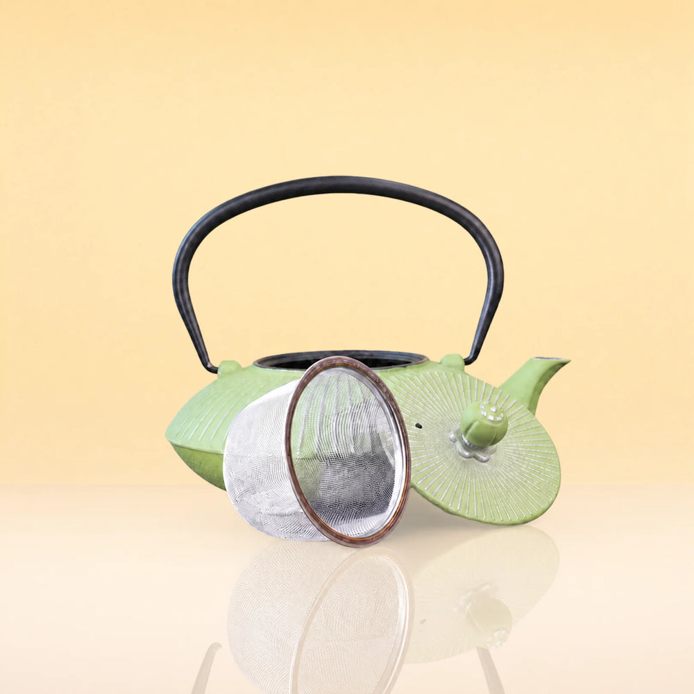 OliveInfusionTeaKettle2 - A light green ceramic tea kettle with a black metal handle, shown with its lid off and a round metal tea infuser resting in front, set against a warm beige gradient background.