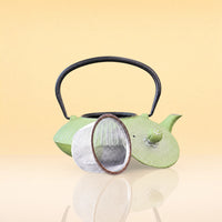 OliveInfusionTeaKettle2 - A light green ceramic tea kettle with a black metal handle, shown with its lid off and a round metal tea infuser resting in front, set against a warm beige gradient background.