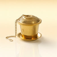 Royal Vault Infuser – Gold-toned stainless steel mesh tea infuser with a domed perforated lid, matching saucer, and a gold chain with hook, photographed on a warm cream background.