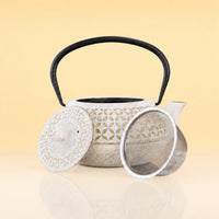 SageTerriorIronTeapot - A ceramic teapot with a textured off-white body, floral cutout pattern, black handle, matching lid, and a separate metal infuser strainer, displayed on a reflective surface against a warm beige background.