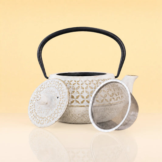 SageTerriorIronTeapot - A ceramic teapot with a textured off-white body, floral cutout pattern, black handle, matching lid, and a separate metal infuser strainer, displayed on a reflective surface against a warm beige background.