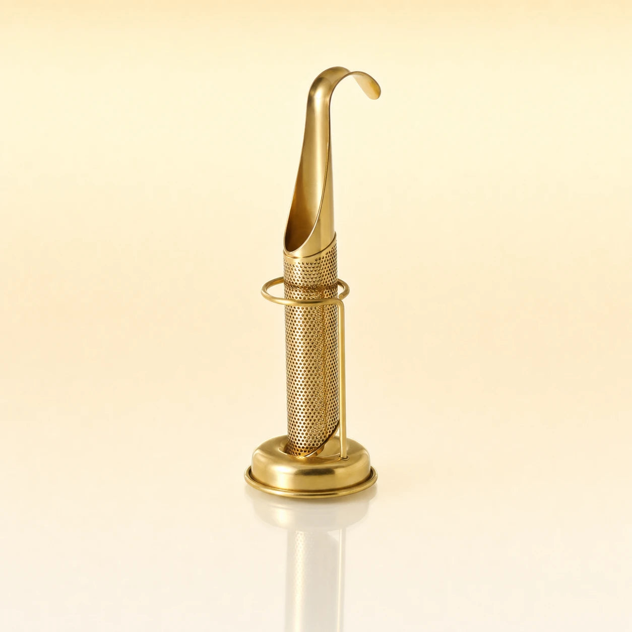 Signature Grip Infuser – Gold-toned stainless steel upright tea infuser with a hook-shaped handle, cylindrical mesh body, and a circular base stand, photographed on a warm cream background with a soft reflection below.