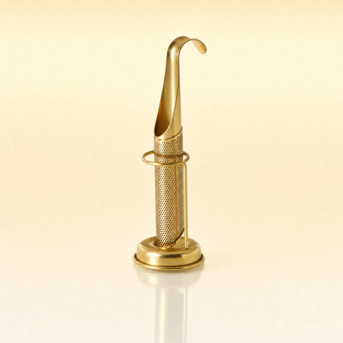 Signature Grip Infuser – Gold-toned stainless steel upright tea infuser with a hook-shaped handle, cylindrical mesh body, and a circular base stand, photographed on a warm cream background with a soft reflection below.