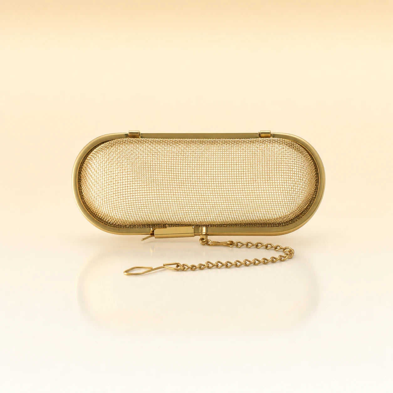 Silk Oval Infuser – Gold-toned stainless steel oval-shaped tea infuser with a hinged lid, rounded edges, and a delicate gold chain with clasp, photographed on a warm cream background with a soft reflection below.