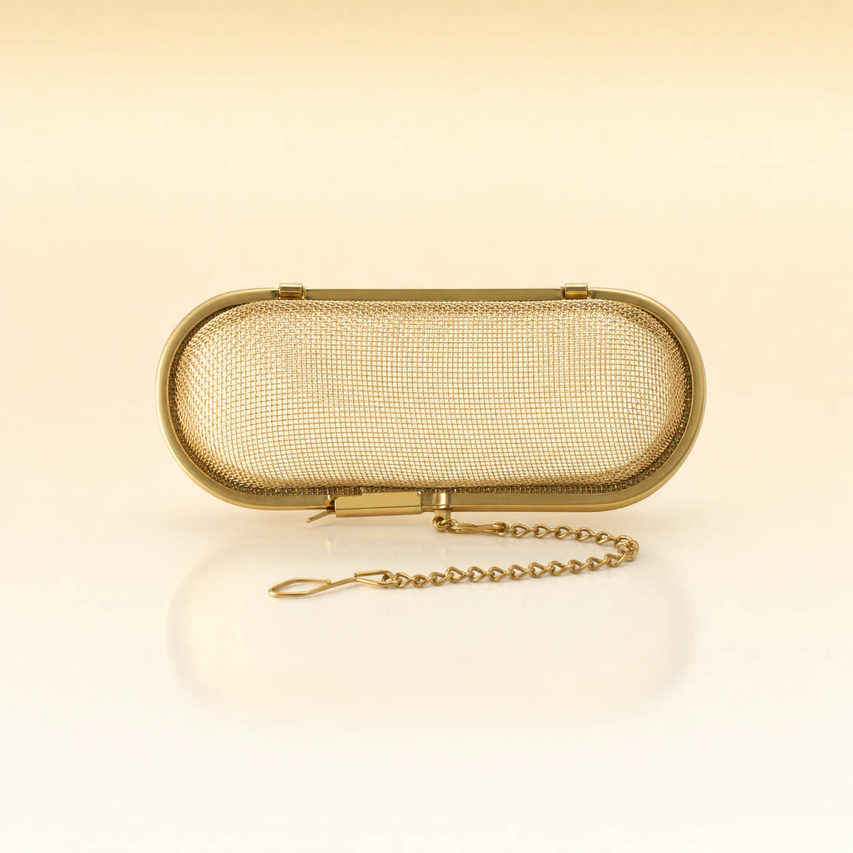 Silk Oval Infuser – Gold-toned stainless steel oval-shaped tea infuser with a hinged lid, rounded edges, and a delicate gold chain with clasp, photographed on a warm cream background with a soft reflection below.