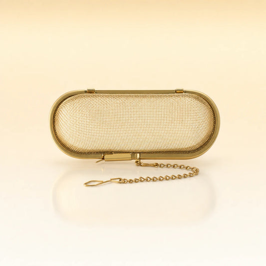 Silk Oval Infuser – Gold-toned stainless steel oval-shaped tea infuser with a hinged lid, rounded edges, and a delicate gold chain with clasp, photographed on a warm cream background with a soft reflection below.