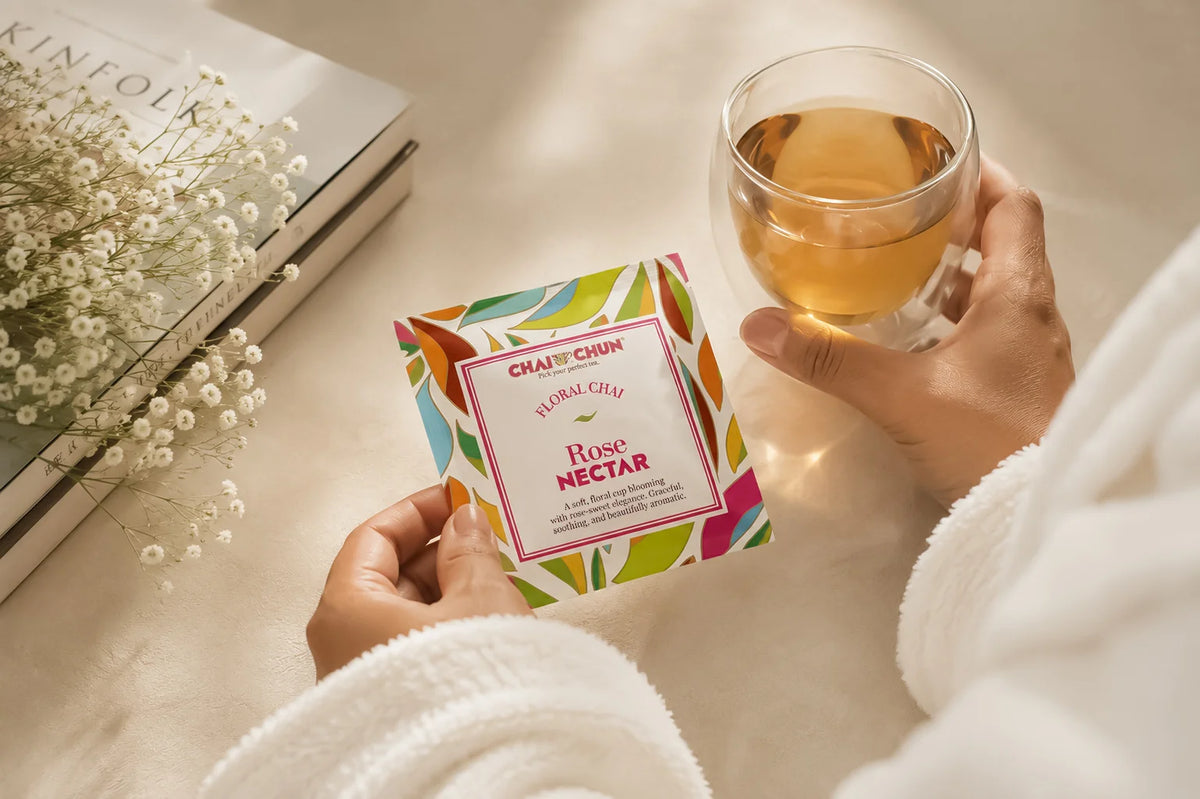 Spa - A person in a white robe holds a tea packet labeled Chai Chai Rose Nectar next to a glass of brewed tea on a warm beige surface with books and baby's breath flowers nearby.