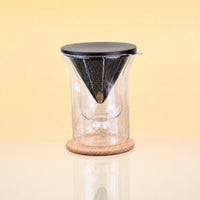TeaChalice-DoubleWalledGlass_TeaInfuserSet - A clear glass pour-over coffee dripper with a black mesh cone filter, a black lid, and a round cork base, shown on a warm beige background.