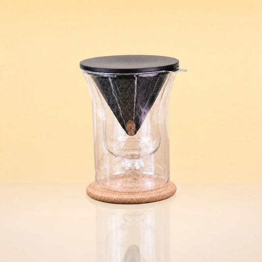 TeaChalice-DoubleWalledGlass_TeaInfuserSet - A clear glass pour-over coffee dripper with a black mesh cone filter, a black lid, and a round cork base, shown on a warm beige background.