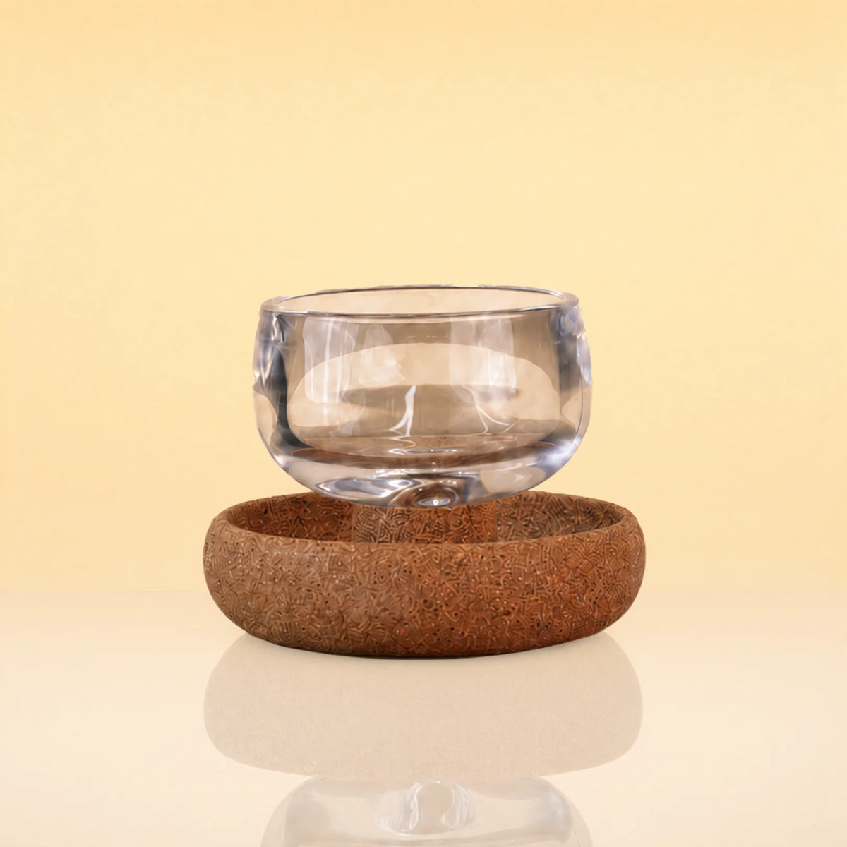TeaOrbitCup - A clear glass cup floating above a round brown cork coaster on a reflective surface, set against a warm beige background.