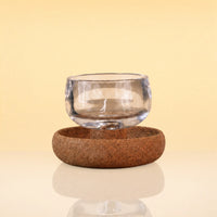 TeaOrbitCup - A clear glass cup floating above a round brown cork coaster on a reflective surface, set against a warm beige background.