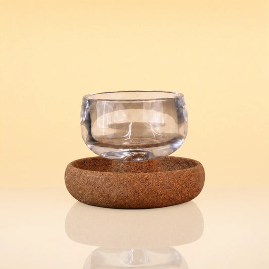 TeaOrbitCup - A clear glass cup floating above a round brown cork coaster on a reflective surface, set against a warm beige background.