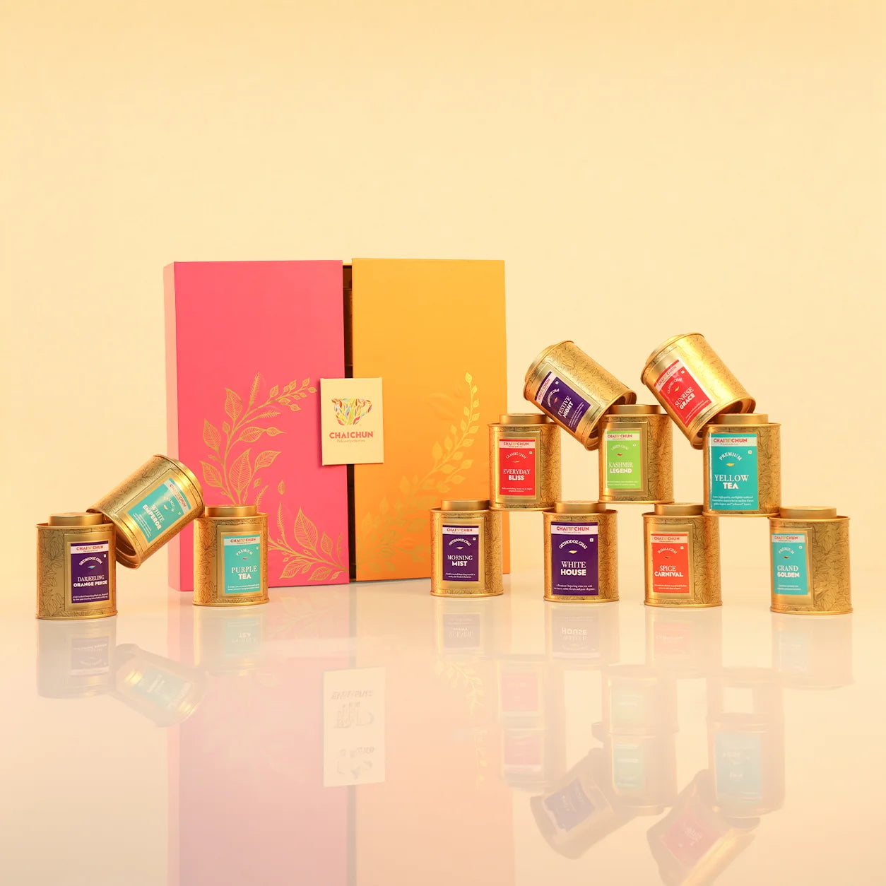Tea_Ritual_Collection - A tea gift set featuring multiple small cylindrical metal tea tins in gold with colorful labels, arranged in front of a pink and orange decorative box. The tins display labels for various tea varieties such as Darjeeling, Assam, and Nilgiri.