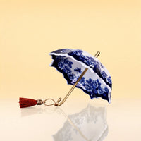 UmbrellaTeaInfuser - A miniature ceramic umbrella with a blue and white floral pattern, a gold-tone handle, and a red tassel, set against a warm beige background with a soft reflection below.