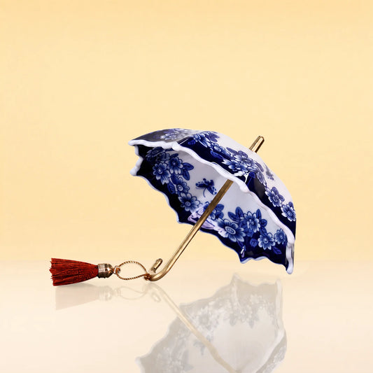 UmbrellaTeaInfuser - A miniature ceramic umbrella with a blue and white floral pattern, a gold-tone handle, and a red tassel, set against a warm beige background with a soft reflection below.