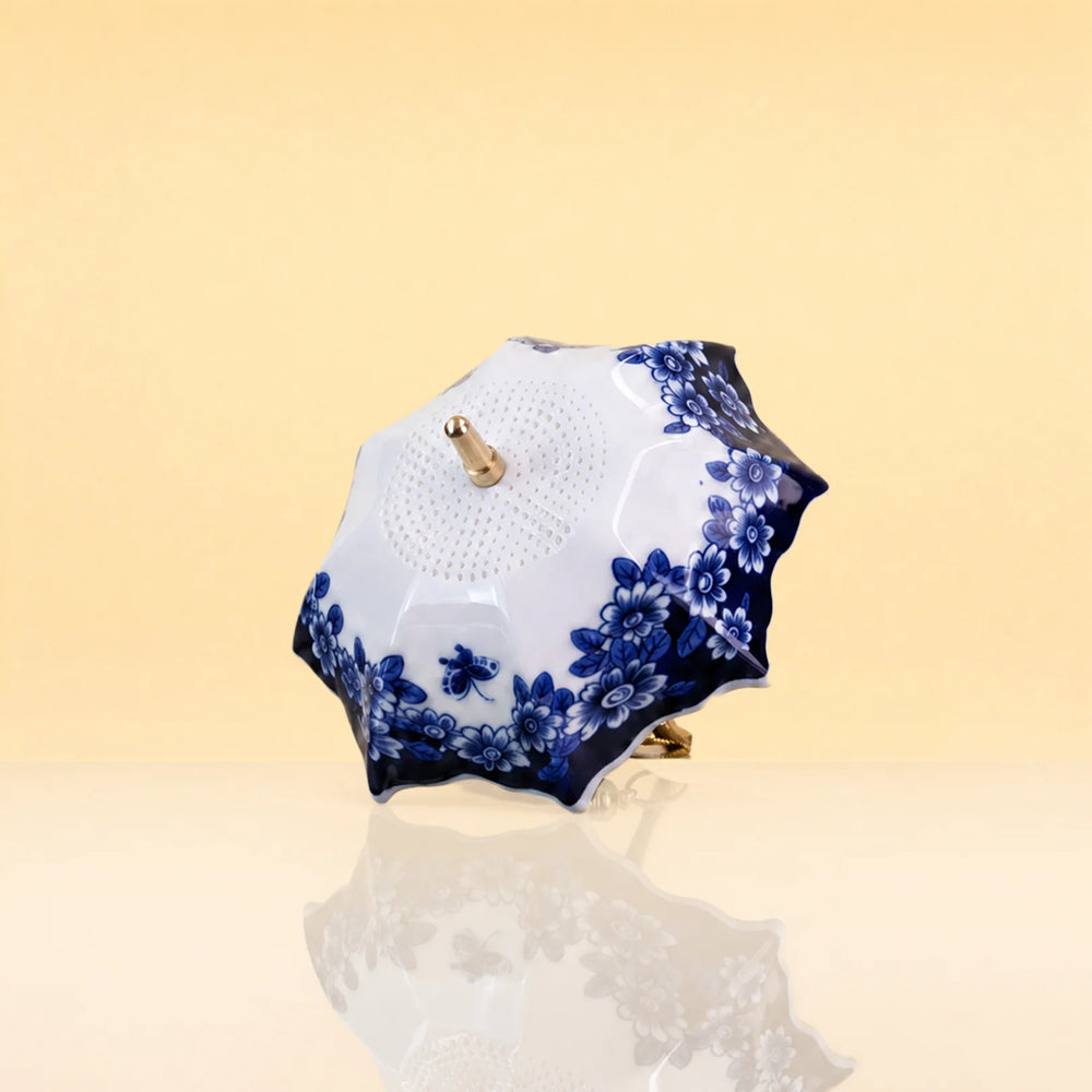 UmbrellaTeaInfuser2 - A small decorative parasol with a white body and blue floral pattern, featuring gold-tone metal hardware, set against a warm beige background with a soft reflection below.