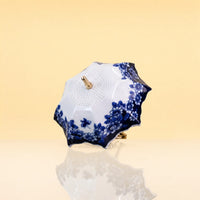 UmbrellaTeaInfuser2 - A small decorative parasol with a white body and blue floral pattern, featuring gold-tone metal hardware, set against a warm beige background with a soft reflection below.