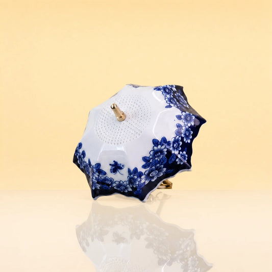 UmbrellaTeaInfuser2 - A small decorative parasol with a white body and blue floral pattern, featuring gold-tone metal hardware, set against a warm beige background with a soft reflection below.