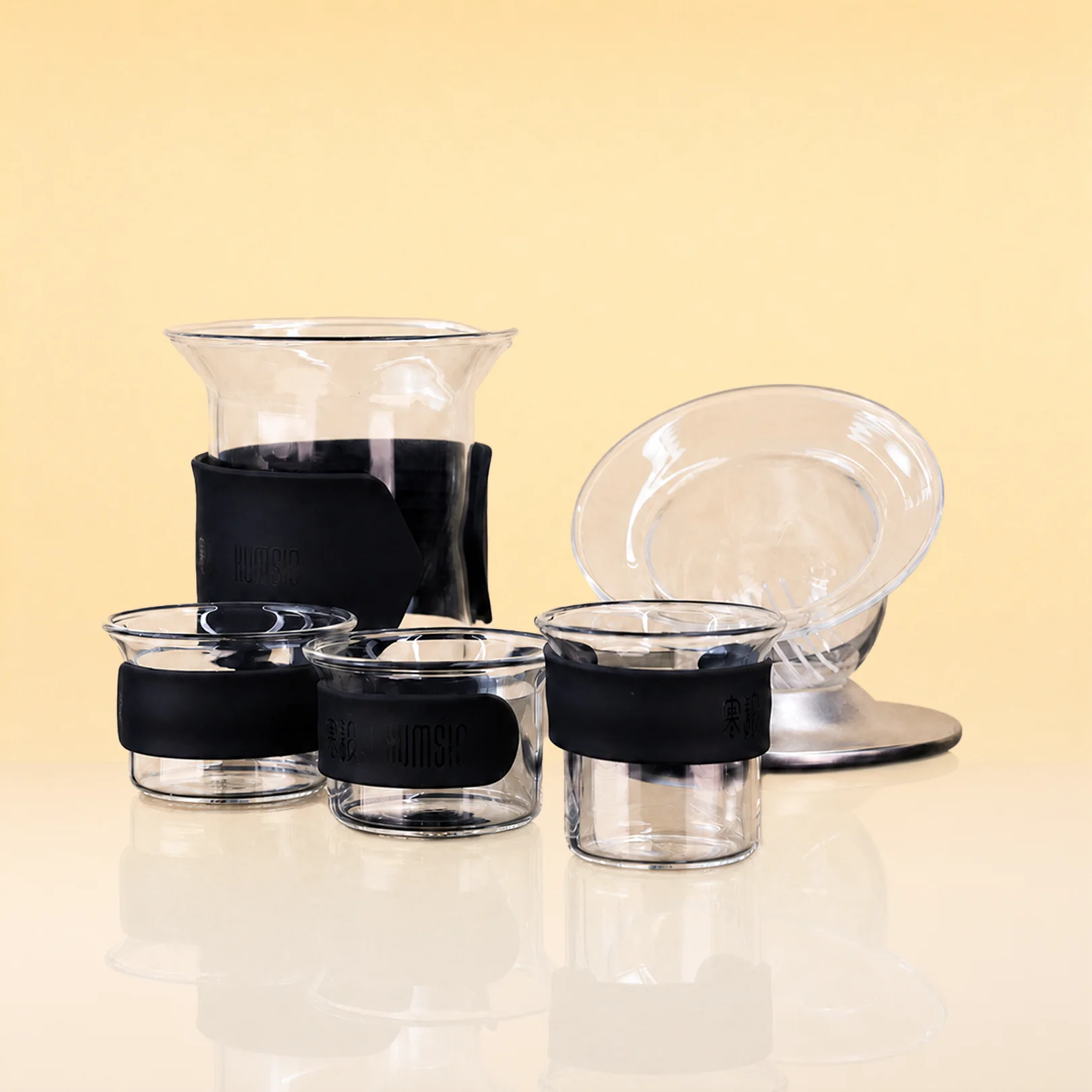 VoyageSet-PortableGlassTeaSet - A set of clear glass pour-over coffee accessories with black silicone sleeves, including a dripper, a small cup, a medium cup, a large carafe, and a small saucer, arranged on a reflective white surface against a warm beige background.