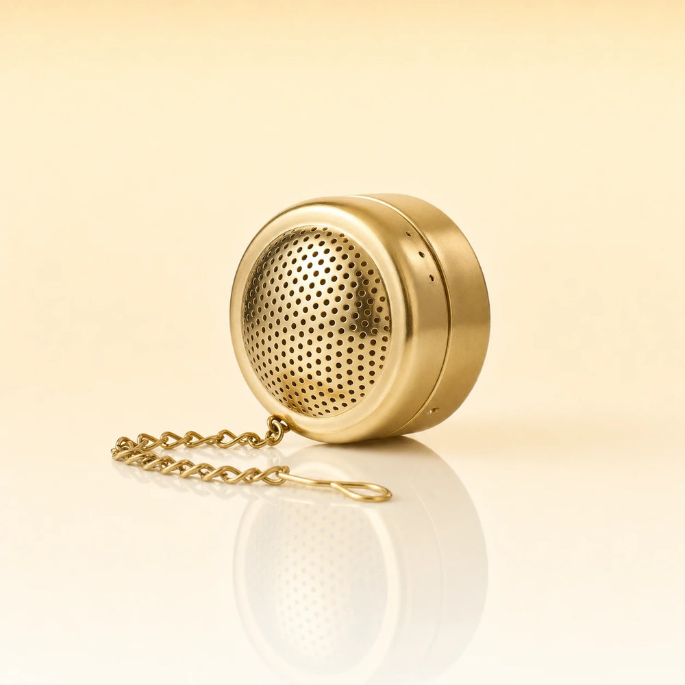 ZEN POD INFUSER - A gold-finished metal tea infuser with a matching chain, shown on a clean, light beige reflective surface.