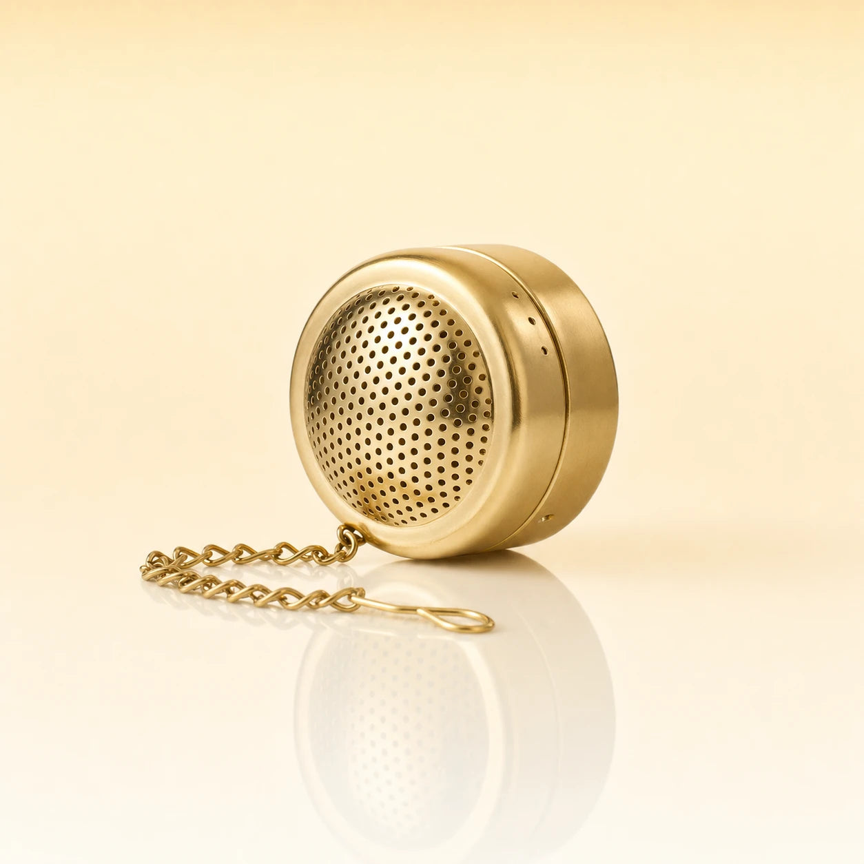ZEN POD INFUSER - A gold-finished metal tea infuser with a matching chain, shown on a clean, light beige reflective surface.
