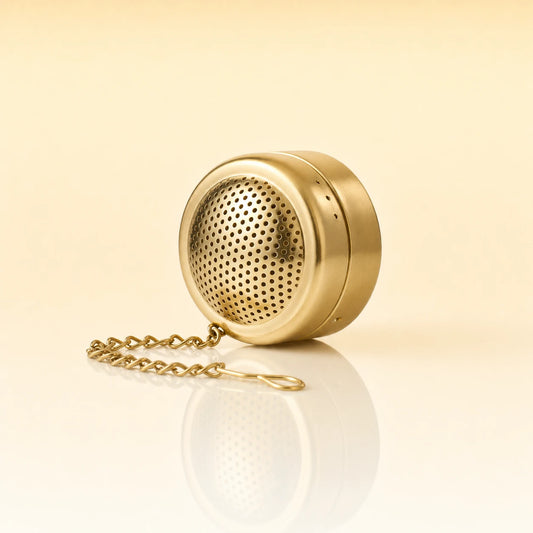 ZEN POD INFUSER - A gold-finished metal tea infuser with a matching chain, shown on a clean, light beige reflective surface.