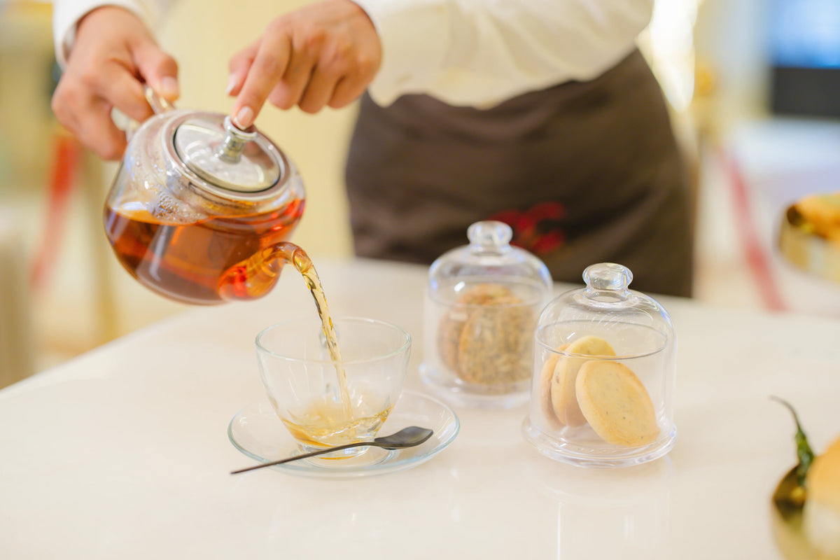 bar-cafe-and-restaurant - A person in a white shirt and brown apron pours amber tea from a clear glass teapot into a glass cup on a saucer. Two glass cloches containing cookies sit on the white table in a softly blurred indoor setting.