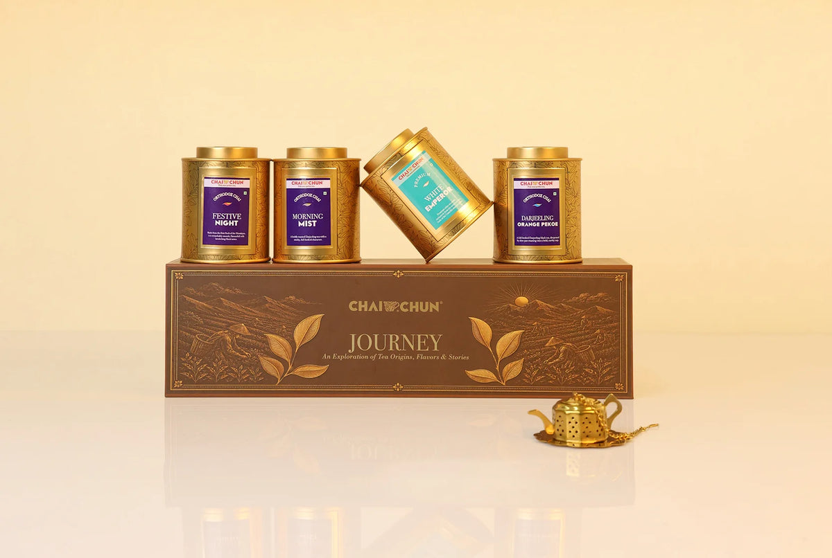 Corporate-gifting - A boxed tea gift set from CHAI+CHUNJ Journey, featuring four gold tea tins with purple and teal labels arranged on top of a brown decorative box, with a small gold teapot on a saucer in front, against a warm beige background.