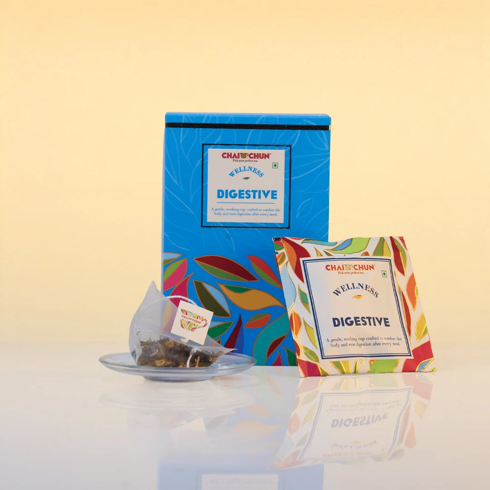 A tea product display showing a blue rectangular box labeled 'CHAITCHUK WELLNESS DIGESTIVE' behind a single tea bag on a small plate and a single sachet with a multicolor leaf pattern and the same label, all on a light beige background.