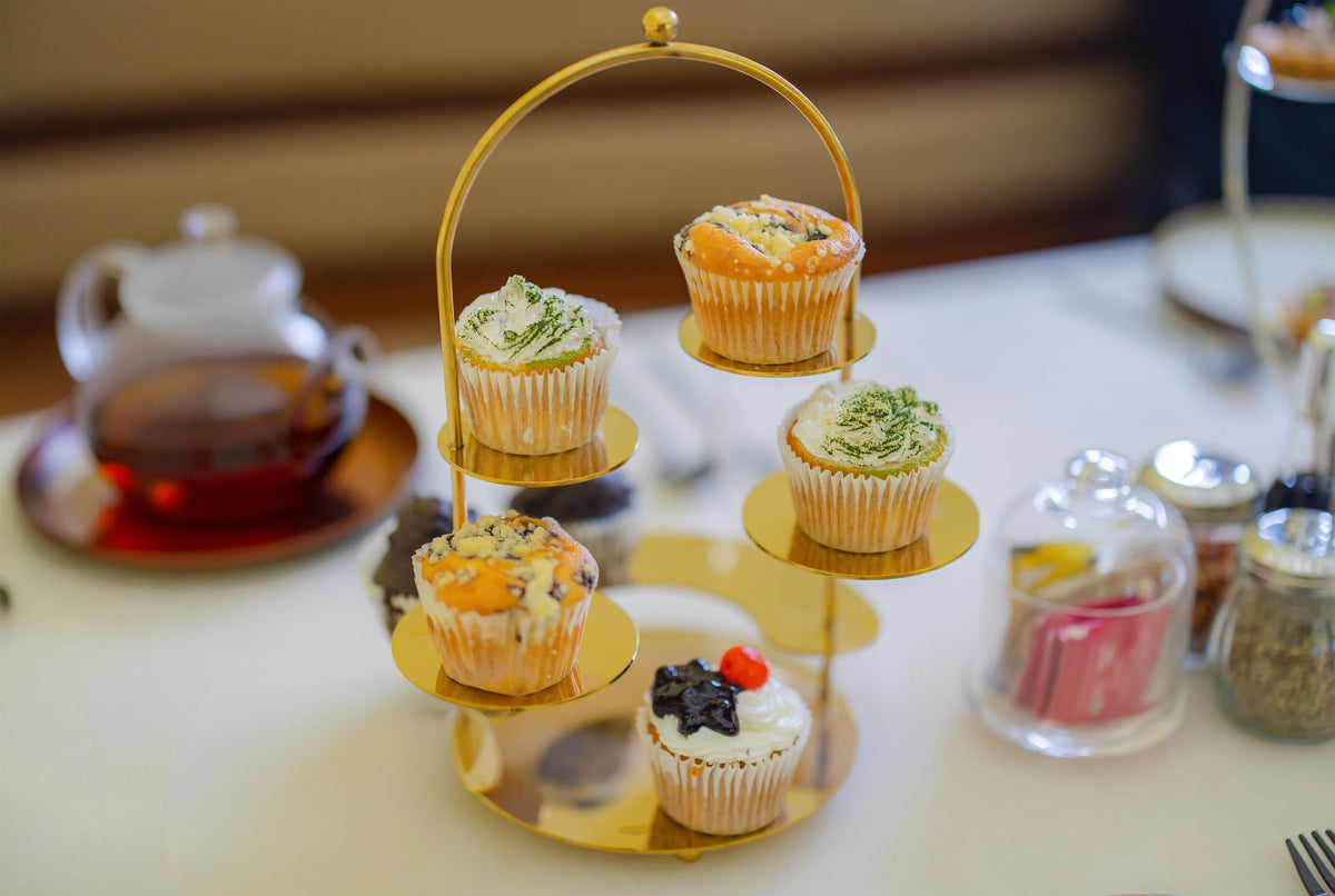 dining-from-am-to-pm - A gold three-tier cupcake stand holding five decorated cupcakes on a white table, with a teapot and glass jars blurred in the background.