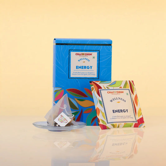 Chaitanya's Wellness & Energy tea set, featuring a blue metal tea tin with colorful leaf patterns, a single tea bag on a small saucer, and an individual tea bag packet in front, all set against a warm beige background.
