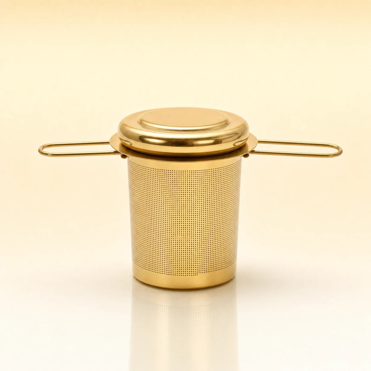 Heritage Cup Infuser – Gold-toned stainless steel cylindrical tea infuser with a flat lid, two wire side handles, and a smooth base, photographed on a warm cream background with a soft reflection below.