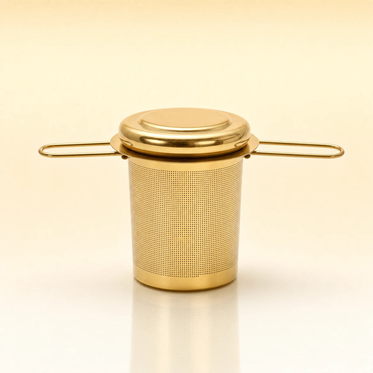 Heritage Cup Infuser – Gold-toned stainless steel cylindrical tea infuser with a flat lid, two wire side handles, and a smooth base, photographed on a warm cream background with a soft reflection below.
