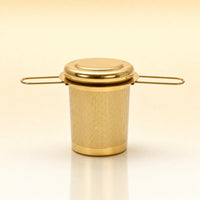 Heritage Cup Infuser – Gold-toned stainless steel cylindrical tea infuser with a flat lid, two wire side handles, and a smooth base, photographed on a warm cream background with a soft reflection below.