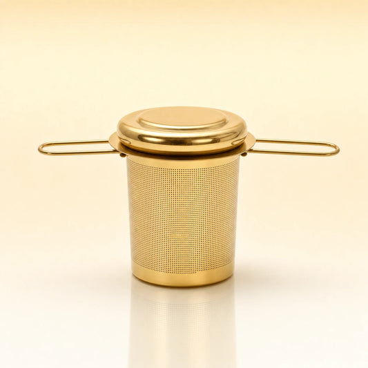 Heritage Cup Infuser – Gold-toned stainless steel cylindrical tea infuser with a flat lid, two wire side handles, and a smooth base, photographed on a warm cream background with a soft reflection below.