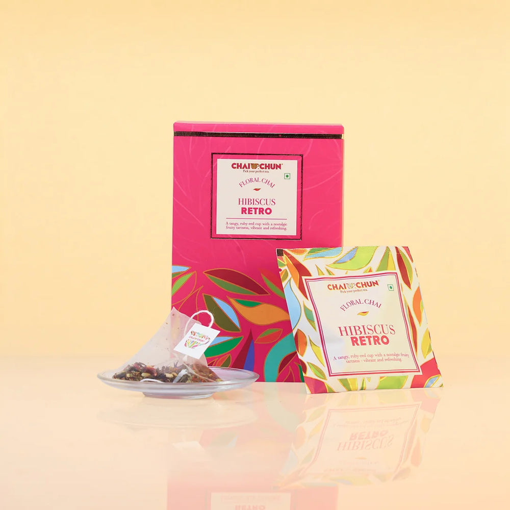 Chaplin Chai Hibiscus Retro tea packaging, featuring a bright pink box with colorful tropical leaf patterns, a single tea bag on a small plate, and an individual tea packet in front, all set against a warm beige background.