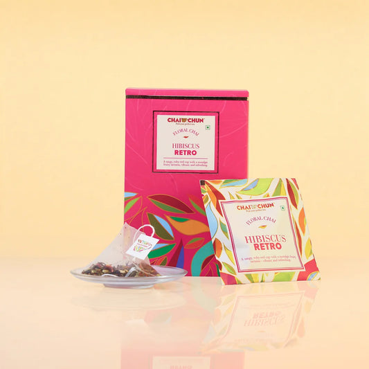 Chaplin Chai Hibiscus Retro tea packaging, featuring a bright pink box with colorful tropical leaf patterns, a single tea bag on a small plate, and an individual tea packet in front, all set against a warm beige background.