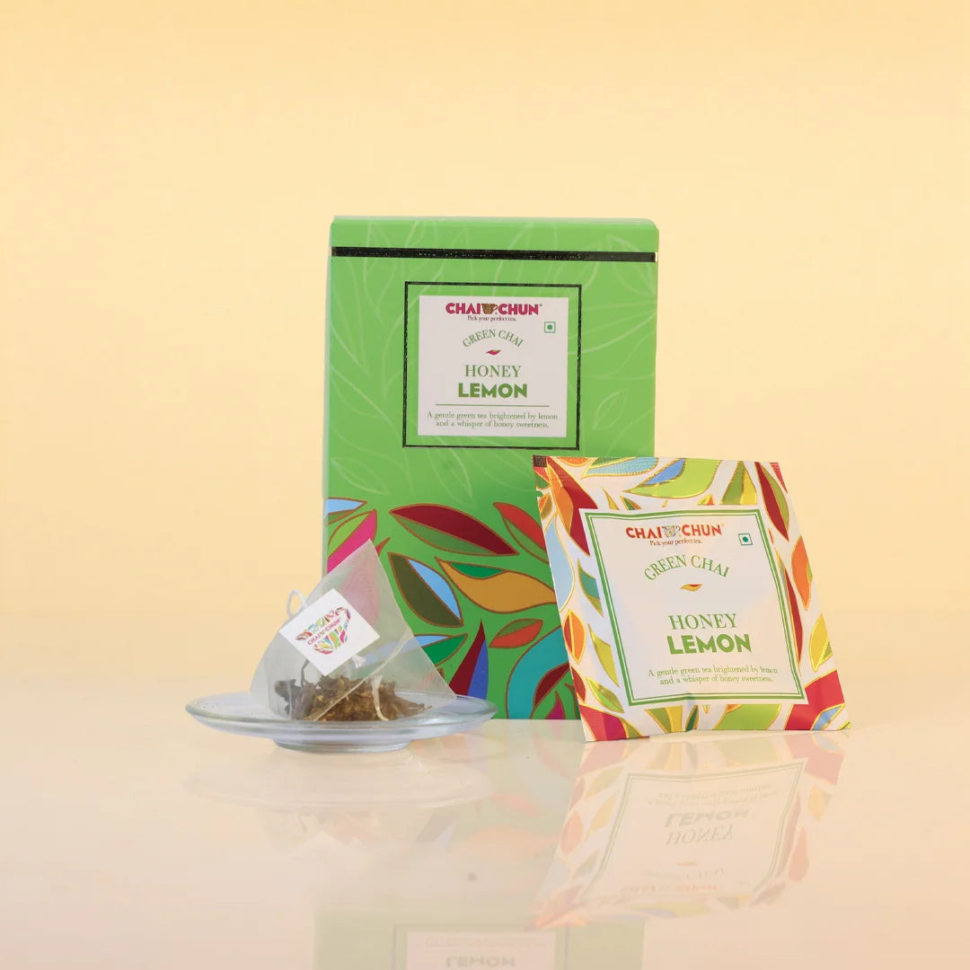 kashmiri-kahwa - Chai Chai Chun Green Chaai Honey Lemon tea set, featuring a green box with colorful leaf patterns, a single tea bag on a small glass saucer, and a single tea bag packet in front, all on a reflective beige surface.