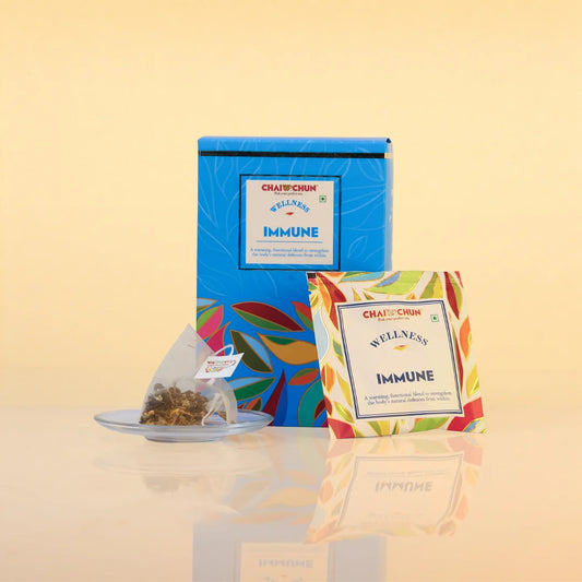 immune_tea_bag - Chai Chaiim Wellness Immune tea set featuring a blue box, a single teabag on a small glass saucer, and a colorful tea bag packet on a beige background.