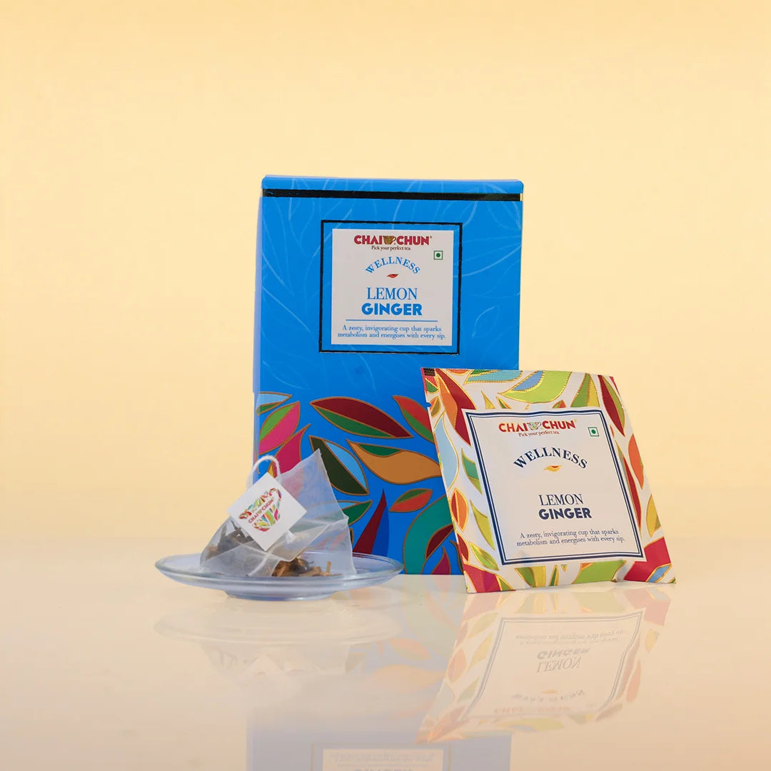 Chai Chai Well Nee Lemon Ginger tea set, featuring a blue box with colorful leaf patterns, a single tea bag on a small saucer, and an individual tea bag packet with a matching multicolor patterned border, all set against a warm beige background.