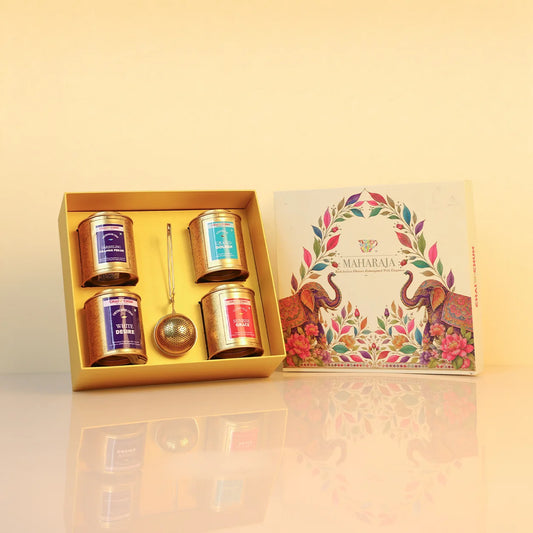maharaja-collection-Vol-1 - A boxed tea gift set displayed on a glossy cream surface against a warm beige background. The open gold tray contains four small labeled tea tins and a metal tea scoop, with the illustrated outer box standing beside it.