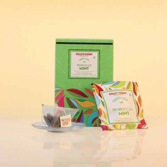 A box of Chai Chai Moroccan Mint Green Chai tea alongside two individual tea bags on a small saucer, set against a warm beige background with a soft reflection below.