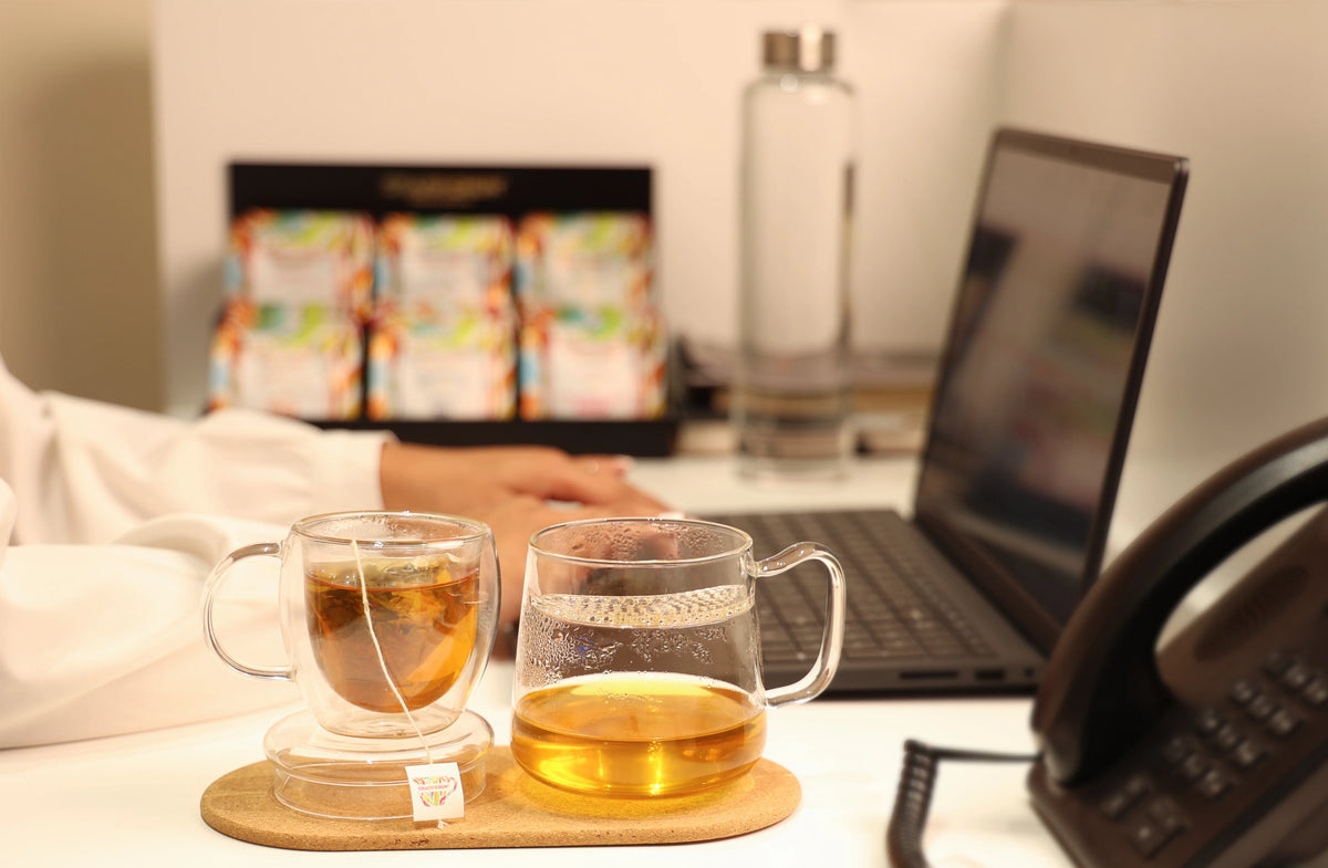 office-scene- Two clear glass cups filled with tea on a wooden tray in the foreground, with a person's hands using a laptop in a warm indoor setting. Tea bag packaging is visible in the blurred background.