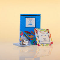 A blue tea box labeled CHAI CHUN Slim & Trim stands on a reflective surface next to a single tea bag on a saucer and a smaller front-facing package card. The background is a soft warm beige gradient.