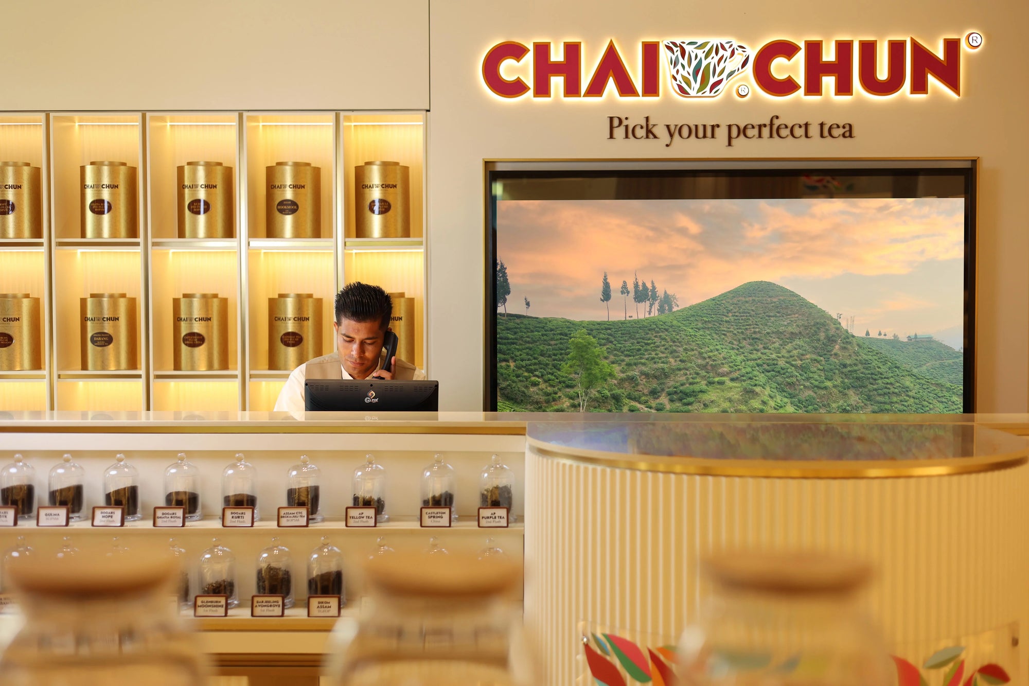 talk-to-us-directly  - A retail tea display area under a large CHAI CHUN sign reading 'Pick your perfect tea.' A man sits behind the counter using a laptop, with shelves of gold tea tins and glass jars on display.